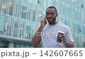 Good morning. African man drinking coffee to go talking on smartphone outdoor. Man with cell phone paper cup walking on street. Man making answering call by cellphone having conversation by mobile 142607665