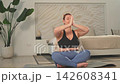 Yoga mindfulness meditation. Woman practicing yoga in living room at home. Plus size female sitting in lotus pose meditating smiling relaxing indoor. Girl doing breathing practice. Yoga at home 142608341