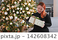 Merry Christmas. Woman unpacking open up gift box near Christmas tree. Girl in living room with Christmas tree holding opening gift box with surprise face. Woman celebrating Christmas eve at home 142608342