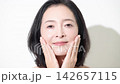 A beauty portrait of a woman in her 50s with beautiful skin, smiling directly at the camera and touching her cheek as she applies skincare products. 142657115