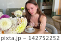 Young woman taking a selfie beside white coffee cup in cafe, adjusting her braided hair and talking to smartphone camera during a casual social media livestream 142752252