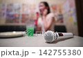 A blurred karaoke room scene with a microphone in the foreground and a woman singing in the background. 142755380