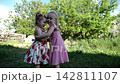 Two cute little sisters hugging each other tightly on green grass outdoor. Beautiful small girls smiling and embracing in summer garden on sunny day. Carefree children show family love and friendship 142811107