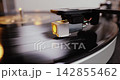 Detailed close-up of a turntable tonearm with white cartridge headshell, positioned over a black vinyl record on the platter, highlighting audio equipment precision. 142855462