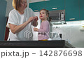 A Caucasian mother looks after her young daughter at home and gently cleans her face while the girl stands on a small stool. The everyday scene represents loving motherhood, childcare, and family car 142876698