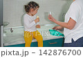 Little girl eats a banana on the kitchen countertop while her mother cleans around her. The scene portrays family life, parenting, household responsibilities, and a warm home environment. Mid shot. 142876700