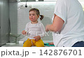 Young mother organizes and cleans the kitchen while her little daughter has a banana on the countertop. The home scene illustrates motherhood, household chores, family comfort, and daily routine. Mid 142876701