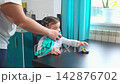 Young mother cleans the kitchen while her little daughter sits at the table nearby. The cozy domestic scene represents motherhood, family life, and everyday household routine. Mid shot. 142876702