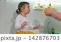 Mother tidies the kitchen as her young daughter enjoys a banana while sitting on the countertop. The cozy domestic scene illustrates motherhood, family bonding, and everyday life. Mid shot. 142876703