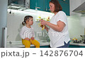 Young mother cleans the kitchen while her little daughter sits on the countertop eating a banana. The scene represents family life, childcare, and everyday home routine. Mid shot. 142876704