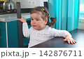 Little Caucasian girl sits at the kitchen table at home. The everyday family scene represents childhood, home life, comfort, and a natural domestic routine 142876711