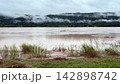 Timelapse video showing the wide brown river flowing past green mountains covered in low thick fog. Beautiful scenic landscape of tropical wilderness on a cloudy rainy day. 142898742
