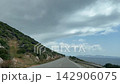 Driving Along a Scenic Coastal Road with Rocky Hillsides and Ocean Views on a Cloudy Day 142906075