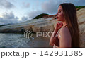 Young woman in swimwear standing on sandy shoreline in profile, gazing toward distant horizon with calm expression beside warm sunset light and eroded sandstone cliffs 142931385
