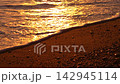 Golden sunset waves gently wash over the sandy pebble shoreline during twilight. Upward tilting camera movement reveals a massive coastal rock silhouette. 142945114