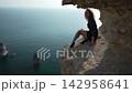 Woman cliff sea, elegant lady sitting on dangerous seaside rock edge looking out over the turquoise ocean abyss during a sunny day. 142958641