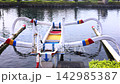 Traditional outrigger canoe floats in still river water, with painted support arms and narrow hull centered in frame. Boat remains docked near landscaped bank, suggesting peaceful transport. 142985387