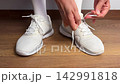 Close up of a woman tying shoelaces on white sneakers indoors. Sport shoes on wooden floor. Preparing for workout, fitness, jogging, training or daily casual activity. 142991818