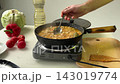 Hand stirring bubbling curry in cast iron skillet on electric burner in bright home kitchen with carrots tomatoes onions nearby, steam rising above saucy curry sauce 143019774