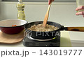 Hands stir meat in cast iron pan on portable stove under bright kitchen light, capturing cooking action, prep mood, and close view of vegetables and cutting board 143019777