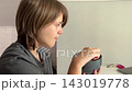 Side profile shows healthcare worker in gray scrubs seated at bright countertop, stirring dark ceramic mug with spoon. Soft natural light on clean interior walls creates relaxed routine mood. 143019778
