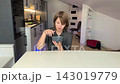 Young child sits at white dining table in bright contemporary kitchen, reaching toward bowl of ice cream and eating slowly. 143019779