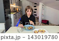 Young woman at kitchen table extends plate of savory pastries toward viewer. Counter, appliances, and neutral walls sit in background as soft warm indoor lighting supports calm inviting mood. 143019780