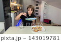 Close view of calm dining moment as woman eats granola with cucumber slices at bright kitchen table, spoon action captured in natural morning daylight with cozy home interior focus on food. 143019781