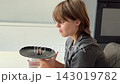 Side profile of woman in gray shirt holding brownie on plate beside glass of water in bright kitchen, relaxed snack moment with soft daylight 143019782