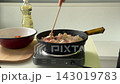 Hands stir meat in cast iron pan on portable stove under bright kitchen light, capturing cooking action, prep mood, and close view of vegetables and cutting board 143019783