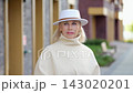 Stylish older woman in white hat smiles outdoors, lifting gloved hand to her brim. This portrait keeps focus on graceful fashion styling and serene expression along modern building columns. 143020201