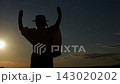 Silhouetted woman lifts both arms toward sunset. Hat tipped profile stays dark against bright horizon, creating serene silhouette as figure moves into triumphant pose with raised hands above head. 143020202
