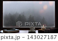 A view of a misty, tranquil forest from a cozy window seat, accompanied by warm coffee, candlelight, and a book. 143027187