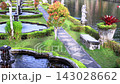 Ornamental garden fountains shimmer beside quiet pond in sunny tropical park. Curved water basins, stone statue, trimmed lawns and patterned walkway create serene landscape with vivid greenery. 143028662
