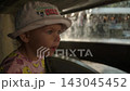 A little girl in a bucket hat and a pink T-shirt happily jumps up and down and waves her arms, looking around at the dense streams of water falling from the fountain and turning her head. 143045452
