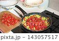 A woman is cooking zucchini caviar by adding diced tomatoes and bell peppers to a pan with fried zucchini, onions, and carrots in a cozy kitchen setting. 143057517