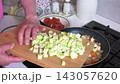 A woman prepares zucchini caviar by adding chopped zucchini to fried onions and carrots. Fresh tomatoes are also present on the table. The cooking takes place in a kitchen. 143057620