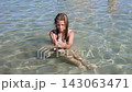 Young girl sitting in clear shallow water and gently moving her hands beneath the surface during a sunny outdoor swim 143063471
