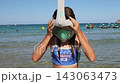 Young girl adjusts full face snorkeling mask while standing in shallow tropical sea with swimmers and boats visible behind her 143063473