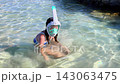 Young girl Wearing Snorkel Mask Sitting and Moving Hands in Clear Shallow Seawater Beside Rocky Shoreline During Outdoor Swimming Activity 143063475