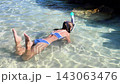 Young girl in blue bikini swimming slowly through clear shallow tropical water while holding a snorkel near a rocky shoreline 143063476