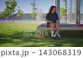 A casual roller skater enjoys a sunny day by the waterfront, preparing for an adventure while taking a break before gliding on the smooth pavement in the park. 143068319