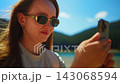 Young woman wearing sunglasses taking a close-up smartphone selfie outdoors near a serene lake with 143068594
