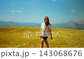 Woman walking through sunny open meadow with distant mountain range, wearing casual summer outfit 143068676