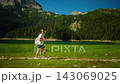 Young woman hiking along a tranquil lakeside trail surrounded by lush green forest and towering 143069025