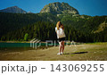 Young woman with backpack walking along tranquil mountain lake shore, surrounded by lush forest and 143069255