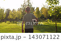 Boy walks across green grass toward sunset, holding soccer ball in one hand. Child is seen from behind in sunny park. Warm golden light highlights outdoor recreation, youth and summer activity. 143071122