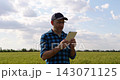 Farmer uses tablet while monitoring wheat crops in field. Smiling agricultural worker combines digital technology with traditional farming. Modern agriculture, smart farming and rural innovation. 143071125