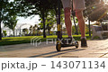 Teenager riding kick scooter through park, close-up of legs in sneakers and shorts at sunset. Active outdoor lifestyle highlights youth mobility, independence, recreation, movement and freedom. 143071134