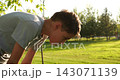 Young boy catches breath after outdoor exercise in sunny park. Tired teenager bends over on green lawn, showing fatigue and recovery. Summer fitness, youth wellness, and active lifestyle concept. 143071139